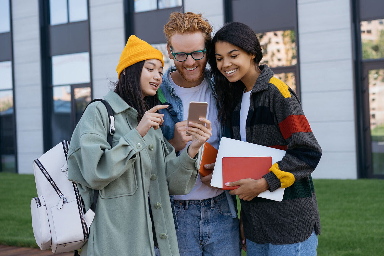 Drei Personen stehen zusammen vor einem modernen Gebäude, lächeln und schauen auf ein Smartphone, das einer von ihnen hält. Eine Person trägt einen grünen Mantel und eine gelbe Mütze, eine andere einen grauen Pullover mit bunten Ärmeln und die dritte Person trägt eine Brille und ein blaues Hemd.